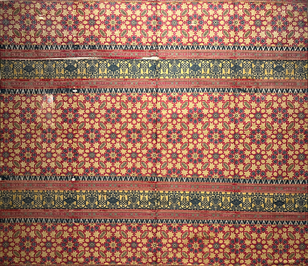 A historic textile features an Islamic pattern of stars and arches in orange, red, yellow and black